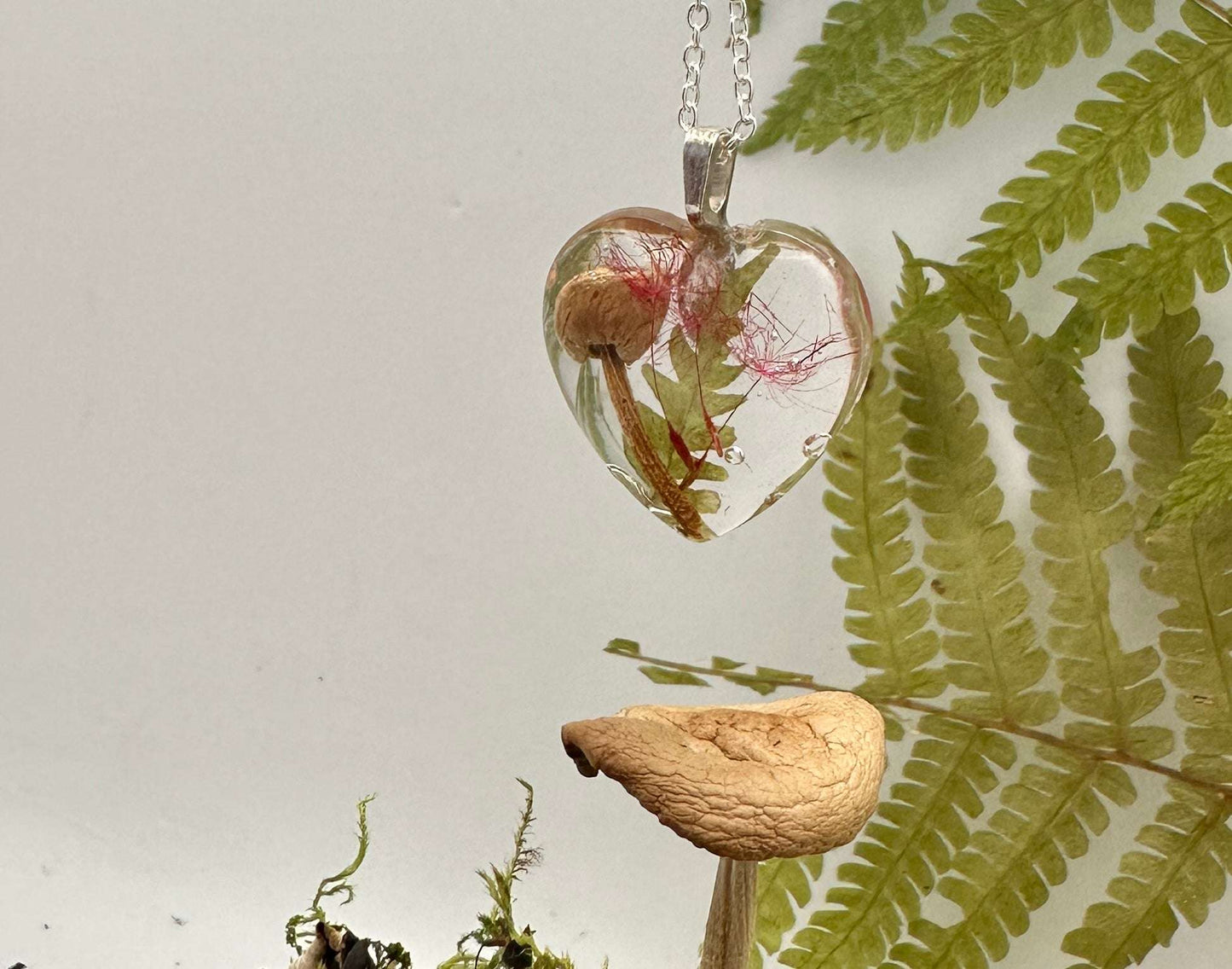 A Resin clear Heart filled with a dried fern, minii mushroom and red tinted dandelion fairy wishes